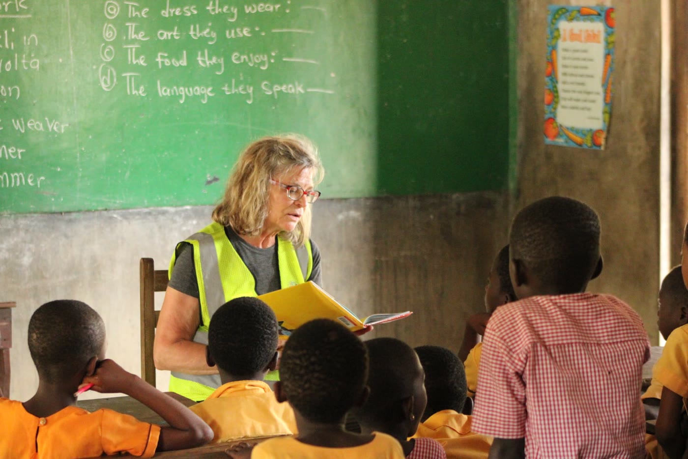 A volunteer seated at the front of a classroom in Ghana, reading from a book to her students